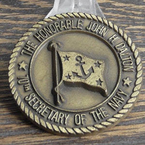 May include: A gold-colored metal coin with a rope-like border. The coin features the text "The Honorable John H Dalton 70th Secretary of the Navy" and a gold-colored flag with a white anchor and stars.