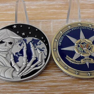 May include: Two circular challenge coins. The left coin features a silver grim reaper holding a globe against a black starry background. The right coin displays a blue center with a white star, the text UNITED STATES INTELLIGENCE COMMUNITY, and a banner with the words COLLABORATUS VIRTUS FIDES.