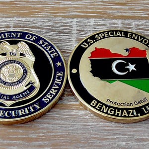 Könnte beinhalten: Zwei runde Herausforderungsmünzen mit goldfarbenen Rändern. Die linke Münze zeigt das Abzeichen des Department of State Diplomatic Security Service. Die rechte Münze zeigt eine Karte von Libyen mit dem Text U.S. SPECIAL ENVOY, Protection Detail, BENGHAZI, LIBYA.