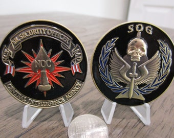 Puede incluir: Dos monedas negras y doradas con diseños intrincados. Una moneda dice "SR. SECURITY OFFICER" y "CENTRAL INTELLIGENCE AGENCY". La otra moneda presenta una calavera y las letras "SOG". Ambas monedas se exhiben en soportes transparentes.