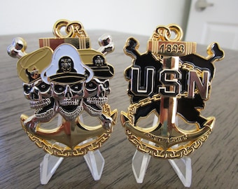 May include: Two gold-colored US Navy challenge coins. The first coin features three skull heads wearing a gold and white sailor hat with the text "USN" on the hat. The second coin features a black and gold anchor with the text "USN" and "1893" above the anchor and "ASK THE CHIEF" below the anchor.