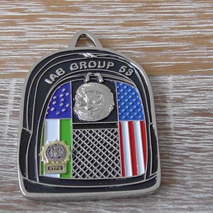 May include: A silver-toned metal challenge coin shaped like a backpack with a black border. The text IAB GROUP 53 is at the top. It features a silver circular emblem, a blue and white star pattern, a red and white striped section, and a gold NYPD badge.