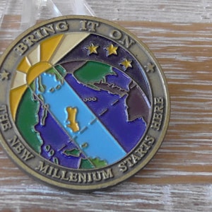 May include: A round, bronze-colored coin with the text "BRING IT ON" and "THE NEW MILLENNIUM STARTS HERE". The coin features a colorful world map design with a rising sun and three stars.
