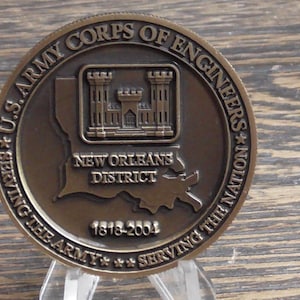 May include: A bronze coin commemorating the U.S. Army Corps of Engineers New Orleans District. The coin features a stylized image of a fort with the text "U.S. Army Corps of Engineers * Serving the Nation" and "New Orleans District 1818-2004".