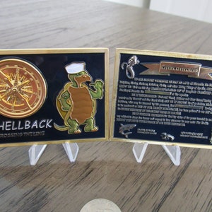 Peut inclure: Une pièce de monnaie en métal doré avec un fond bleu et un motif de boussole d'un côté. L'autre côté représente une tortue de dessin animé avec un chapeau de marin et le texte "Shellback Crossing the Line" et "Imprimis Lege Regis".