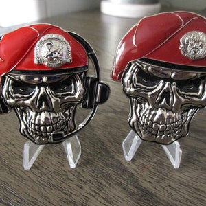 May include: Two silver skull challenge coins with red berets and military insignia. The coins are on clear acrylic stands.