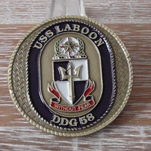 May include: A gold-colored challenge coin with a navy blue background. The coin features the text "USS LABOON DDG 58" and a crest with a trident and the words "Without Fear".