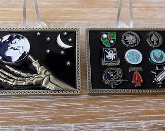 Puede incluir: Dos vitrinas rectangulares con pines metálicos. La vitrina izquierda muestra una mano esquelética que alcanza un globo terráqueo con una luna creciente y estrellas. La vitrina derecha contiene ocho emblemas e insignias de estilo militar.