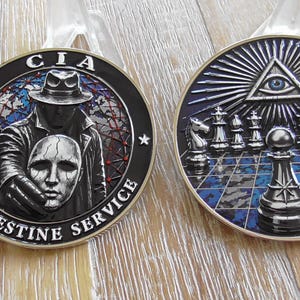 May include: Two circular metal challenge coins with detailed engravings. The left coin features a figure in a trench coat and hat holding a mask with the text CIA CLANDESTINE SERVICE. The right coin displays a chessboard with pieces and an eye.