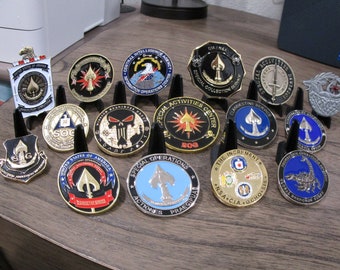 Op de afbeelding: Een verzameling van vijftien ronde en schildvormige uitdagingsmunten met diverse emblemen van militaire en inlichtingendiensten, waaronder schoppensymbolen, adelaars en teksten zoals Central Intelligence Agency en Special Activities Center.