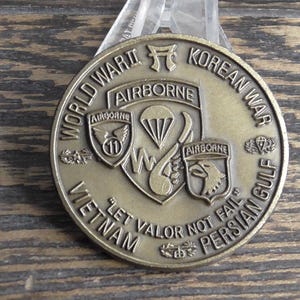 May include: A bronze-coloured commemorative coin with text including "World War II," "Korean War," "Vietnam," and "Persian Gulf." The coin features the word "Airborne" and military emblems. The phrase "Let Valor Not Fail" is also inscribed.