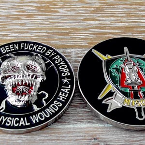 May include: Two circular metal challenge coins with black enamel backgrounds. The left coin features a silver skull with red accents and the text YOU'VE JUST BEEN FUCKED BY PSYOPS BECAUSE PHYSICAL WOUNDS HEAL. The right coin displays a shield emblem with a sword, a green globe, and the text MISOC.
