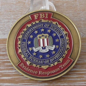 Può includere: Una moneta sfida circolare in metallo con il sigillo dell'FBI in blu, rosso e oro. Il testo recita FBI, Department of Justice, Federal Bureau of Investigation e Evidence Response Team, con le parole Fidelity, Bravery e Integrity.