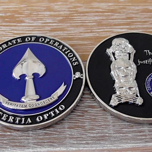 Puede incluir: Dos monedas de desafío metálicas circulares. La moneda de la izquierda presenta un centro azul con una punta de lanza plateada, el texto "DIRECTORATE OF OPERATIONS" y "TERTIA OPTIO", y "VERITATEM COGNOSCERE". La moneda de la derecha es negra con una pila de calaveras plateadas y el texto "The end justifies the means".