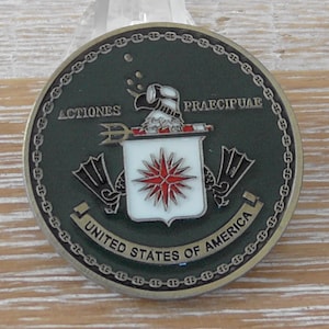 May include: A circular challenge coin with a dark green background and a metallic border featuring a chain link pattern. The center displays a white shield with a red compass rose, flanked by two black swim fins and the text ACTIONES PRAECIPUAE.