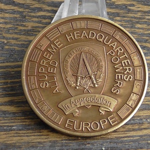 以下が含まれることがあります： 縁に「SUPREME HEADQUARTERS ALLIED POWERS EUROPE」の文字が入った、ブロンズ色の記念コイン。 コインの中央には、交差した剣と「In Appreciation」の文字が入ったエンブレムがあります。 コインは木製の表面に表示されています。