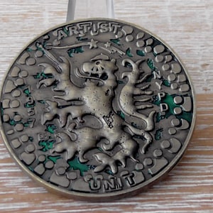 Op de afbeelding: Een ronde bronzen metalen medaille met een centraal reliëf van een leeuw. De tekst ARTIST staat bovenaan, UNIT onderaan en NYPD verticaal aan de rechterkant. Het getal 1957 staat links. De achtergrond is voorzien van groen emaille.