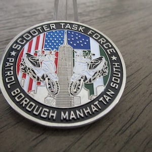 May include: A silver-colored challenge coin with a black border and a white background. The coin features a depiction of two scooters with riders, the Empire State Building, and the American and Italian flags. The text "Scooter Task Force Patrol Borough Manhattan South" is inscribed around the edge of the coin.