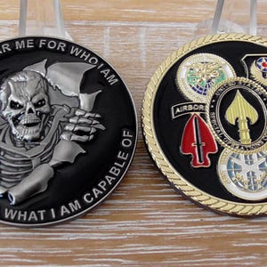 May include: Two circular challenge coins. The left coin is black with a silver skeleton graphic and the text 'DON'T FEAR ME FOR WHO I AM FEAR ME FOR WHAT I AM CAPABLE OF'. The right coin features a gold rope edge and multiple military emblems.