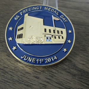 Peut inclure: Une pièce de monnaie dorée avec un fond bleu. La pièce représente un bâtiment avec un mât et un drapeau américain. Le texte "66 Precinct Medal Day June 11th 2014" est inscrit autour du bâtiment.