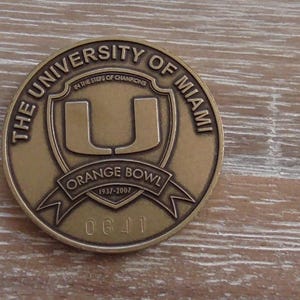 May include: A bronze-colored commemorative coin featuring the University of Miami logo, a stylized 'U' within a shield. The coin is inscribed with "THE UNIVERSITY OF MIAMI" and "ORANGE BOWL 1937-2007". The serial number "0641" is also visible.