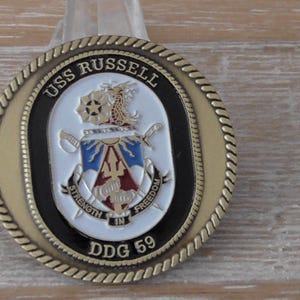 Könnte beinhalten: Eine goldfarbene Münze mit schwarzem Rand und seilartigem Rand. Die Münze trägt den Text "USS RUSSELL DDG 59" und ein zentrales Emblem mit einem Schild, Schwertern und den Worten "STRENGTH IN FREEDOM".