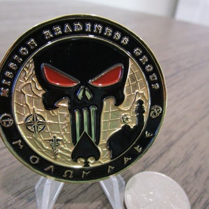 Op de afbeelding: Een ronde gouden munt met een zwarte schedel en rode ogen. De schedel is gecentreerd op een gouden wereldkaart. De munt heeft de tekst "Mission Readiness Group" rond de rand.