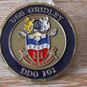 Puede incluir: Una moneda de color dorado con un escudo azul y blanco que presenta un tridente y el texto "IGNIS UBI PARATUS" y "DDG 101". La moneda también tiene el texto "USS GRIDLEY" alrededor del borde.