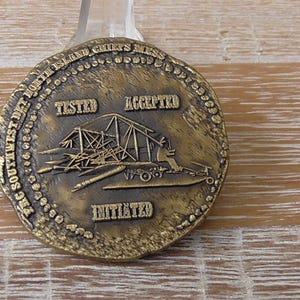 May include: A bronze-colored coin with raised text and imagery. The coin features the words "TESTED", "ACCEPTED", and "INITIATED". A detailed illustration of a structure is in the center. The coin is round with a textured edge.