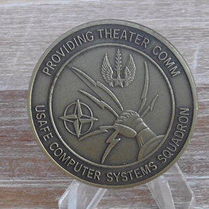 May include: A bronze coin with the words "Providing Theater Comm USAFE Computer Systems Squadron" around the edge. The center of the coin features a stylized lightning bolt and a winged star.