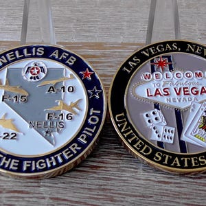 May include: Two circular metal challenge coins. The left coin features a map of Nevada with F-15, A-10, F-16, and F-22 aircraft, labelled 'NELLIS AFB HOME OF THE FIGHTER PILOT'. The right coin displays the 'WELCOME TO FABULOUS LAS VEGAS' sign, dice, and playing cards.