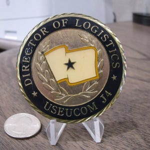 Op de afbeelding: Een gouden munt met een zwarte rand en de tekst "DIRECTOR OF LOGISTICS USEUCOM J4" rond de rand. Het midden van de munt heeft een gouden krans met een witte vlag met een zwarte ster in het midden.
