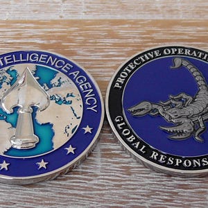 Puede incluir: Dos monedas de desafío metálicas circulares. La moneda de la izquierda presenta un emblema de pica plateado sobre un mapa mundial azul y plateado con el texto CENTRAL INTELLIGENCE AGENCY. La moneda de la derecha muestra un escorpión plateado sobre fondo azul con el texto PROTECTIVE OPERATIONS DIVISION GLOBAL RESPONSE STAFF.