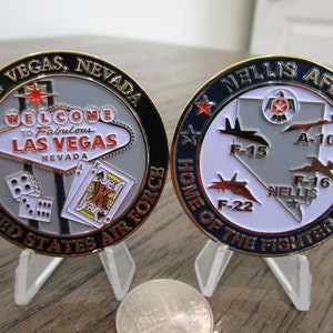 May include: Two gold-coloured challenge coins. The front of the coin features a welcome to Las Vegas sign with dice and playing cards. The back of the coin features a map of Nevada with the text "Nellis AFB Home of the Fighter Pilot" and various fighter jets.