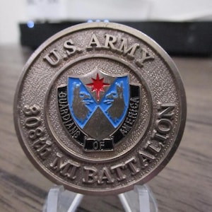 Może przedstawiać: Srebrna moneta armii amerykańskiej z napisem "308th MI Battalion" i "Guardians of America" wokół krawędzi. W centrum monety znajduje się tarcza z czerwoną gwiazdą i dwoma głowami wilków.