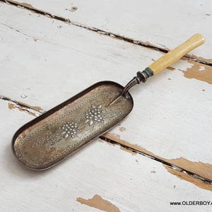 May include: An antique silver-toned crumb scraper with a cream-colored handle. The rectangular tray features a floral design. The handle is attached with a decorative metal piece. The scraper rests on a distressed white wooden surface.