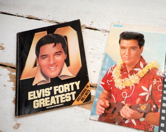 Könnte beinhalten: Zwei Vintage-Vinyl-Schallplatten mit Elvis Presley. Das erste Album ist schwarz mit weißem Text, der "Elvis' Forty Greatest" und "Including 18 No. 1 Hits" lautet. Das zweite Album ist ein farbenfrohes Bild von Elvis Presley, der ein rot-weißes Hawaiihemd mit einer Blumenkette trägt. Der Text auf dem Album lautet "Elvis" und "Blue Hawaii" mit einem rot-weißen Rand.