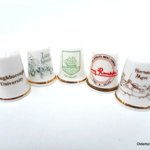 May include: Five white ceramic thimbles with gold trim and various designs. The thimbles feature the names "Loughborough University", "Isea Garden", "Portmadoc", "Ramdden's Fish & Chips", and "Hornsea Mere".