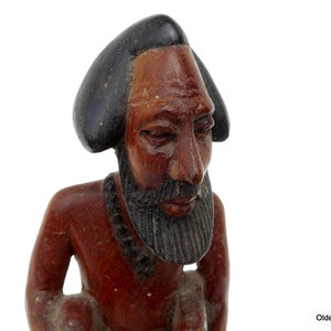 May include: Hand-carved wooden figurine of a person with a dark brown skin tone, wearing a black hat and a beaded necklace. The figurine has a detailed face with a beard and is looking down.