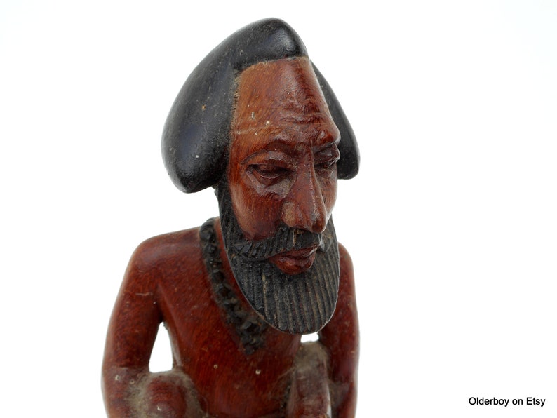 May include: Hand-carved wooden figurine of a person with a dark brown skin tone, wearing a black hat and a beaded necklace. The figurine has a detailed face with a beard and is looking down.