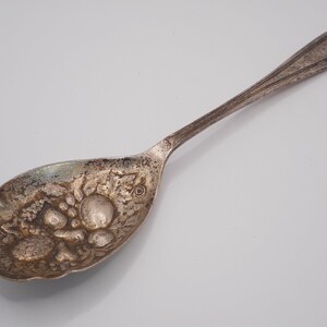 以下が含まれることがあります： ボウルに果物と花の装飾が施された銀のスプーン。ハンドルはシンプルで丸みを帯びた端になっています。