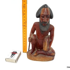 May include: A wooden sculpture of a seated man with a beard and a hat. The sculpture is painted in shades of brown and black.