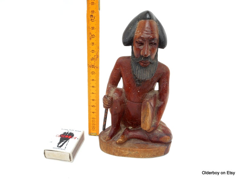 May include: A wooden sculpture of a seated man with a beard and a hat. The sculpture is painted in shades of brown and black.