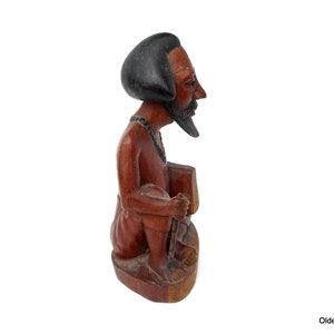 May include: A wooden sculpture of a person sitting with a black hat and beard. The sculpture is carved in a reddish-brown color.