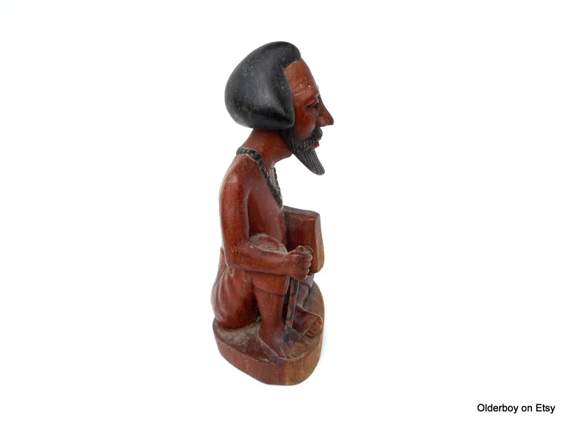 May include: A wooden sculpture of a person sitting with a black hat and beard. The sculpture is carved in a reddish-brown color.