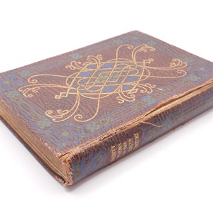 K&ouml;nnte beinhalten: Ein Vintage-Buch mit braunem Lederbezug und goldenen Verzierungen. Der Einband zeigt ein kompliziertes Design mit einem blauen und goldenen Zentrum. Der Titel des Buches lautet "Heilige des Jahrhunderts".