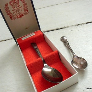 May include: Silver-plated spoon from The Queen's Silver Jubilee 1977, presented in a box with red velvet lining. The box has a navy blue exterior and a printed design with a crown and the text "1977 The Queen's Silver Jubilee". A second spoon is also visible.