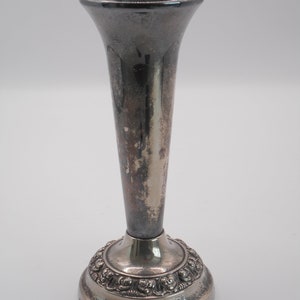 Könnte beinhalten: Eine silberfarbene Blumenvase mit ausgestelltem Oberteil und rundem Sockel. Der Sockel weist ein verziertes Blumendesign auf. Die Vase hat einen schlanken, länglichen Körper und weist Alters- und Gebrauchsspuren auf.