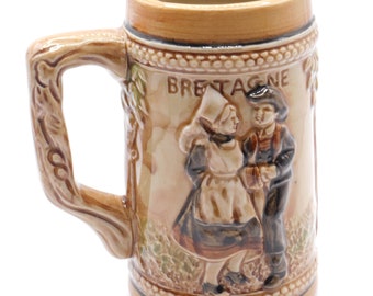 Op de afbeelding: Een bruine keramische bierkan met een reliëfdesign van een stel in traditionele Bretonse kleding. De kan heeft de tekst "BRETAGNE" erop.