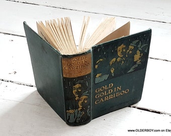 Könnte beinhalten: Ein Vintage-Hardcover-Buch mit dem Titel "Gold Gold in Cariboo" von Clive Phillips-Wolley. Das Buch hat einen dunkelgrünen Einband mit goldenen Lettern und Illustrationen von Personen im Buch.